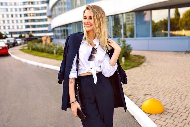 Stilvolle junge Frau, die den trendigen Marineanzug trägt und in der Nähe von modernen Gebäuden, modischen Accessoires, lächelndem Ende posiert, das freien freien sonnigen Sommertag genießt, das nahe Büro geht.