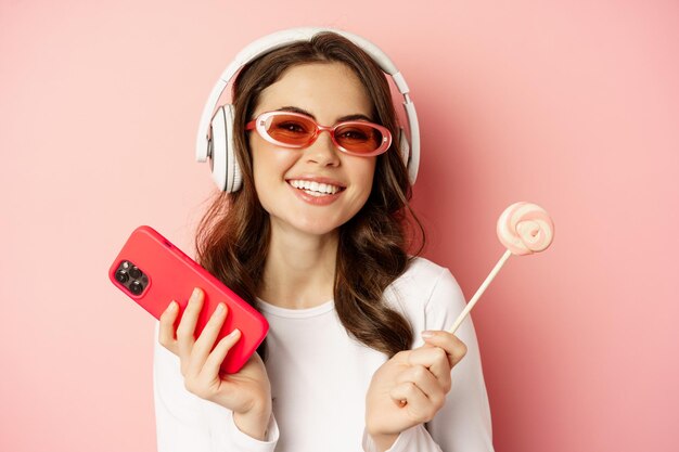 Stilvolle junge Frau mit Lolipop und Handy mit Sonnenbrille und Kopfhörern, die Musiksta...