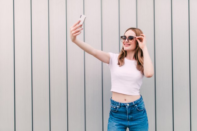 Stilvolle junge Frau nehmen Selfie, das auf weißer Wand aufwirft