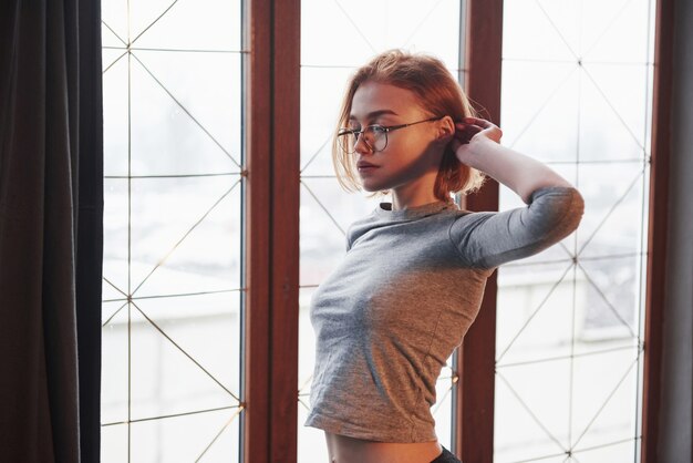 Stilvolle junge Frau. Sexy blondes Mädchen in Brille, Unterwäsche und ohne BH unter dem Hemd posiert in der Nähe des Fensters im Zimmer