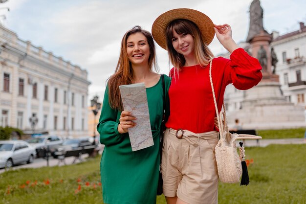 Stilvolle junge Frauen, die zusammen in Europa reisen, gekleidet in frühlingshaftem Outfit und Accessoires, lächeln glückliche Freunde, die Spaß haben, eine Karte zu halten