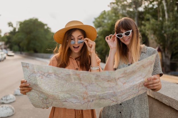 Kostenloses Foto stilvolle junge frauen, die zusammen reisen, gekleidet in trendigen frühlingskleidern und accessoires, die spaß haben, fotos mit der kamera zu machen, die karte hält