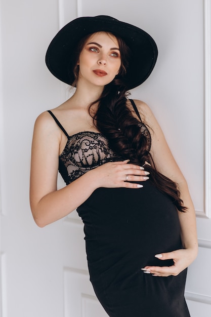 Stilvolle junge schwangere Frau wirft im schwarzen Kleid und im Hut im weißen Studio auf