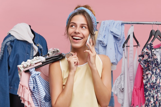 Stilvolle junge weibliche Shopaholic, die auf Handy mit ihrer Freundin spricht und sich über ihre Einkäufe beim Einkaufen im Einkaufszentrum rühmt und am Gestell voller bunter trendiger Kleidungsstücke steht