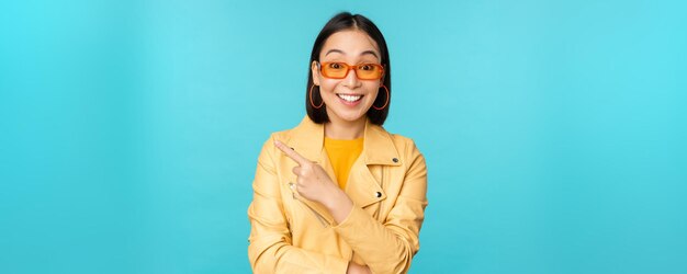 Stilvolle lächelnde asiatische Frau mit Sonnenbrille, die mit dem Finger nach links zeigt und Werbebanner zeigt, die im trendigen gelben Mantel über blauem Hintergrund stehen