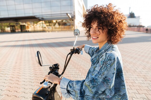 Stilvolle lächelnde Frau, die draußen auf einem modernen Motorrad sitzt