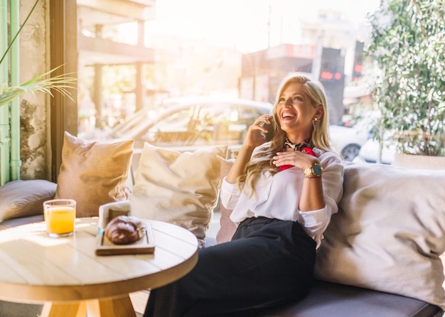 Stilvolle lächelnde junge Frau, die über dem Sofa spricht am Handy im Restaurant sitzt