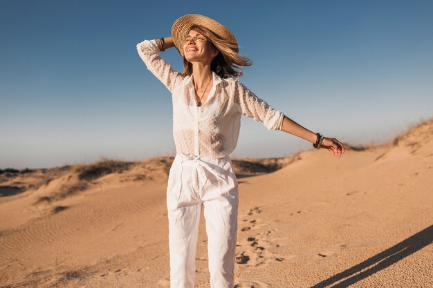 Stilvolle lächelnde schöne glückliche Frau läuft und springt in Wüstensand im weißen Outfit, das Strohhut auf Sonnenuntergang trägt