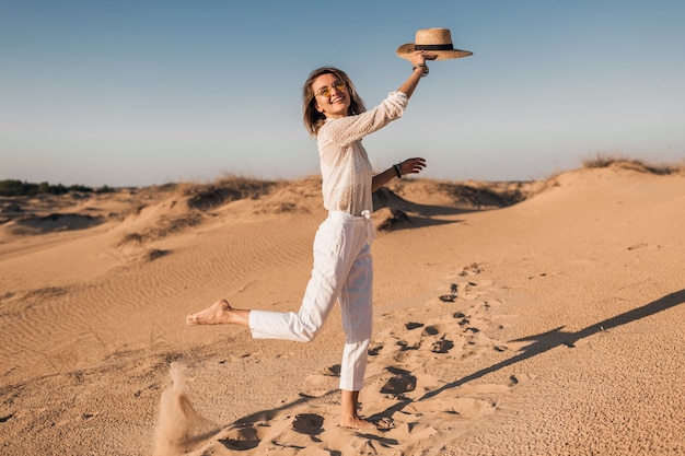 Stilvolle lächelnde schöne glückliche Frau läuft und springt in Wüstensand im weißen Outfit, das Strohhut auf Sonnenuntergang trägt