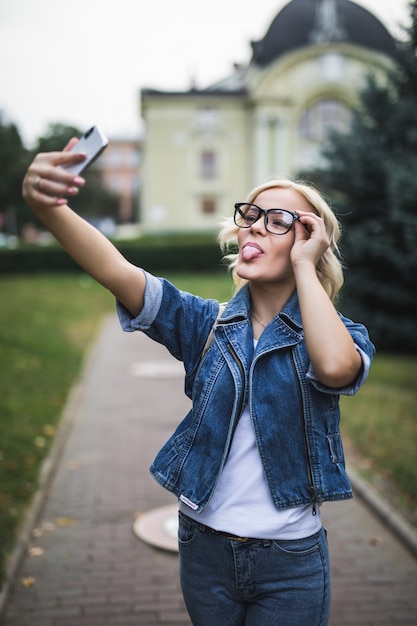 Stilvolle Mode blondes Mädchen Frau in Jeans Suite und Brille macht Selfie auf ihrem Handy in der Stadt am Morgen