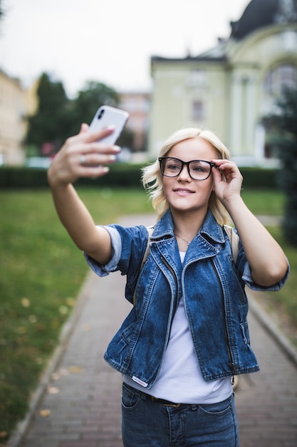 Kostenloses Foto stilvolle mode blondes mädchen frau in jeans suite und brille macht selfie auf ihrem handy in der stadt am morgen