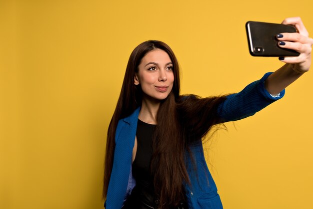 Stilvolle moderne Frau mit langen dunklen Haaren, die blaue Jacke tragen, die selfie mit Smartphone macht