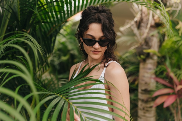 Stilvolle moderne junge frau mit blonder lockiger frisur, die sommerkleidung trägt, die unter tropen im sonnigen sommertag geht. außenfoto des glücklichen lächelnden mädchens hat spaß und genießt wochenende