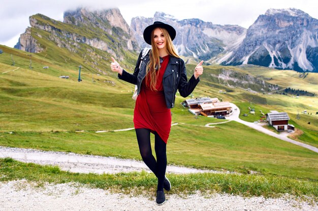 Stilvolle prächtige Frau mit langen Haaren, trendigem Hut, Minikleid und Lederjacke in den Alpen