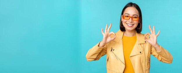 Stilvolle, schöne asiatische Frau mit Sonnenbrille, die erstaunt lächelt und ein okayzeichen zeigt, das etw su empfiehlt