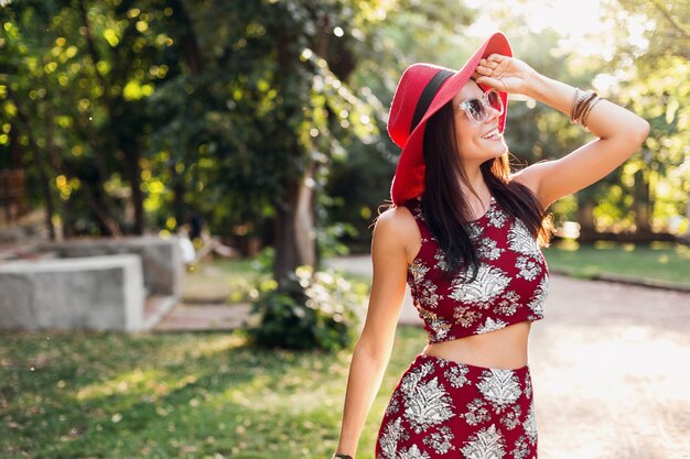 Stilvolle schöne Frau, die im Park im tropischen Outfit geht. Dame im Streetstyle-Sommermodetrend. trägt roten Hut, Sonnenbrille, Accessoires. Mädchen lächelt in fröhlicher Stimmung im Urlaub.