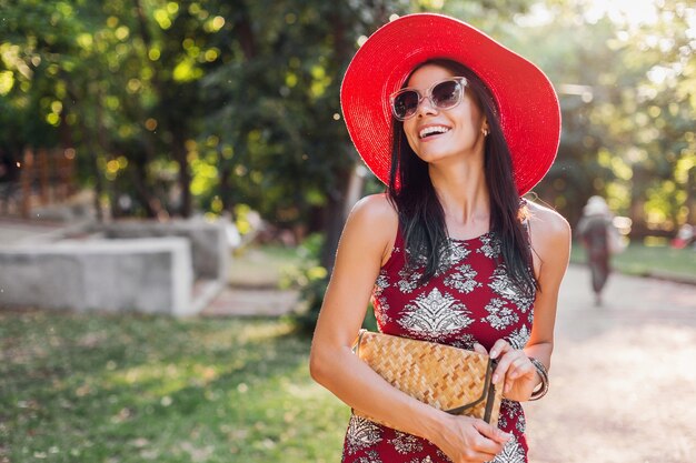 Stilvolle schöne Frau, die im Park im tropischen Outfit geht. Dame im Streetstyle-Sommermodetrend. trägt Strohhandtasche, roten Hut, Sonnenbrille, Accessoires. Mädchen lächelt in fröhlicher Stimmung im Urlaub.