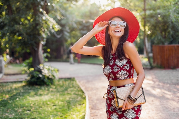 Stilvolle schöne Frau, die im Park im tropischen Outfit geht. Dame im Streetstyle-Sommermodetrend. trägt Strohhandtasche, roten Hut, Sonnenbrille, Accessoires. Mädchen lächelt in fröhlicher Stimmung im Urlaub.