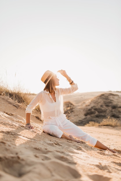 Stilvolle schöne Frau im Wüstensand im weißen Outfit, das Strohhut auf Sonnenuntergang trägt