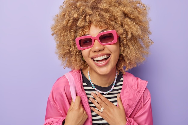 Stilvolle schöne junge Frau mit lockigem, buschigem Haar hält die Hand auf der Brust und lächelt breit trägt eine trendige Sonnenbrille und eine rosafarbene Windjacke, die über violettem Hintergrund isoliert ist Konzept positiver Emotionen