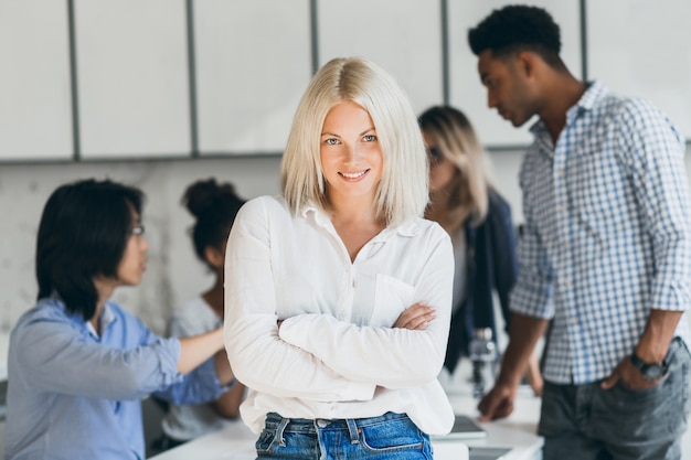 Kostenloses Foto stilvolle sekretärin, die in der selbstbewussten haltung im konferenzsaal steht und lächelt. innenporträt des hübschen blonden büroangestellten, der auf verhandlung mit partnern wartet.