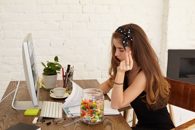 Stilvolle Studentin der Wirtschaftsschule, die am Diplomprojekt arbeitet, an ihrem Arbeitsplatz zu Hause mit Computer, Papierbögen und inneren Gegenständen auf dem Tisch sitzt und Süßigkeiten aus dem Glas isst