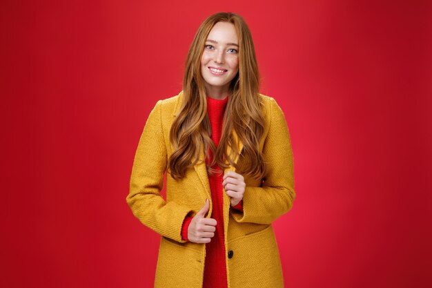Stilvolle und süße, gut aussehende rothaarige Frau in gelbem Mantel auf dem Weg zur Arbeit, Kaffeebestellung mit süßem, freundlichem Lächeln, das den Knopf berührt und auf rotem Hintergrund posiert.