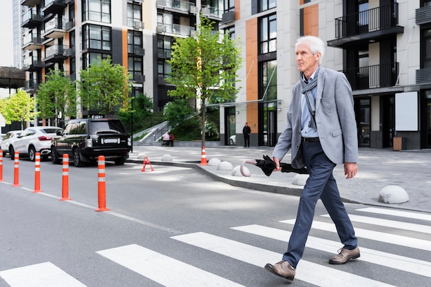 Stilvoller älterer Mann in der Stadt, der die Straße überquert, während er einen Regenschirm hält
