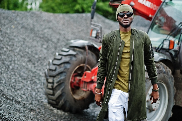 Stilvoller afrikanisch-amerikanischer Mann mit Hut und Sonnenbrille posierte im Freien im Regen gegen einen Traktor mit einem Eimer