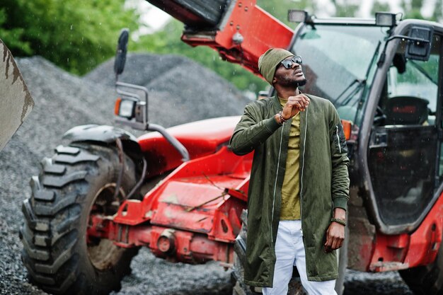 Stilvoller afrikanisch-amerikanischer Mann mit Hut und Sonnenbrille posierte im Freien im Regen gegen einen Traktor mit einem Eimer