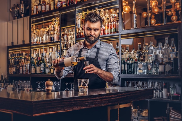 Stilvoller brutaler Barkeeper in Hemd und Schürze macht einen Cocktail im Hintergrund der Bartheke.