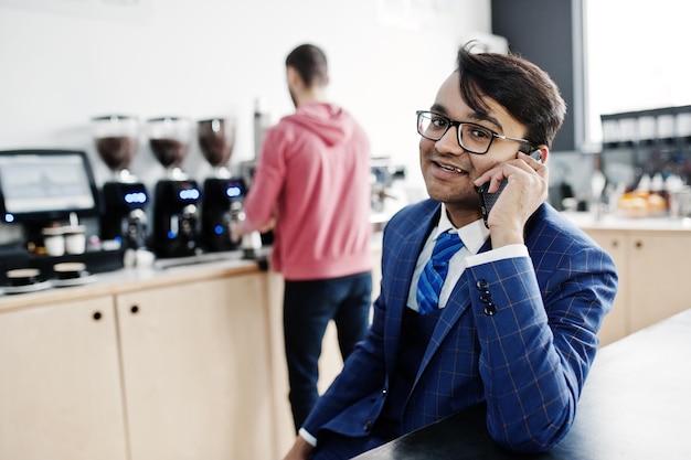 Stilvoller indischer Geschäftsmann in Anzug und Brille, der im Café sitzt und telefoniert