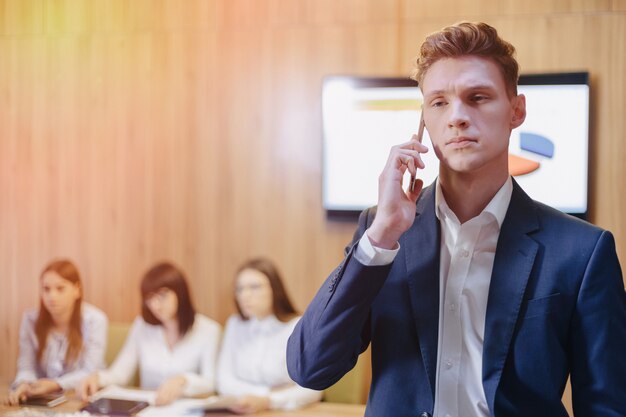 Stilvoller junger Geschäftsmann, der eine Jacke und ein Hemd auf dem Hintergrund eines Arbeitsbüros mit Leuten trägt, die auf einem Mobiltelefon sprechen