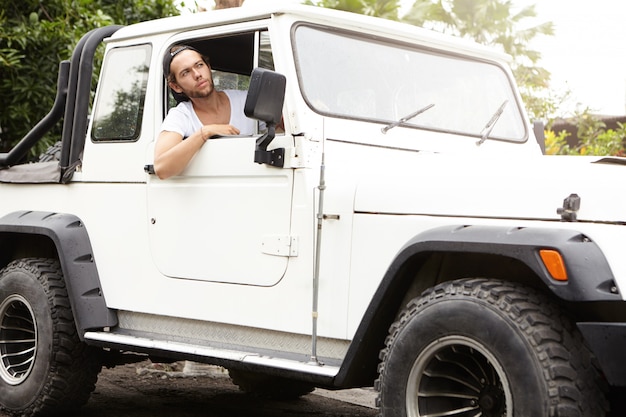 Stilvoller junger kaukasischer Mann, der offenes Fenster seines weißen Sport Utility Vehicle schaut. Unrasierter Mann mit Baseballkappe, der seinen Jeep rückwärts fährt und den Roadtrip genießt