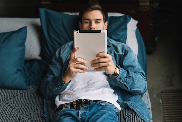 Stilvoller junger Mann, der auf dem Bett betrachtet digitale Tablette liegt