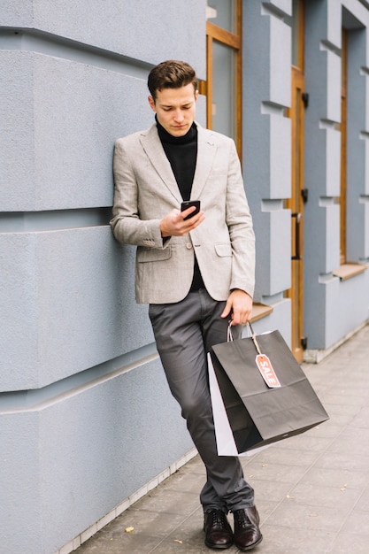 Stilvoller junger Mann, der auf der Wand betrachtet den Smartphone hält Einkaufstaschen sich lehnt