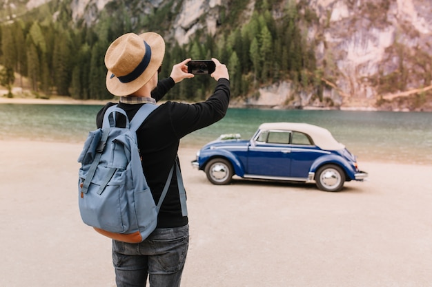 Stilvoller junger Mann, der trendigen Hut trägt, der Spaß am Seeufer hat und Foto der Landschaft macht, die Handy hält