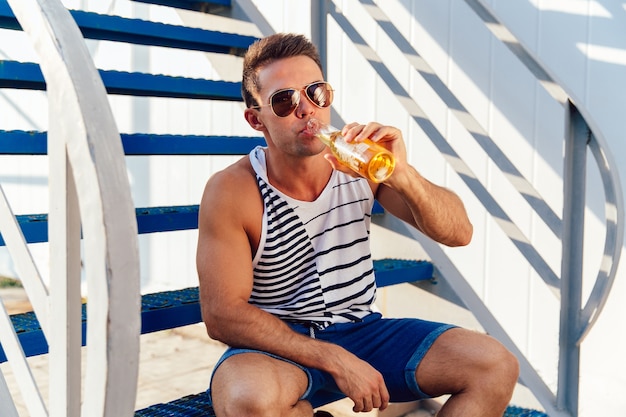Stilvoller junger Mann in der Sonnenbrille ein kaltes Bier beim Sitzen auf Treppe am sonnigen Tag trinkend