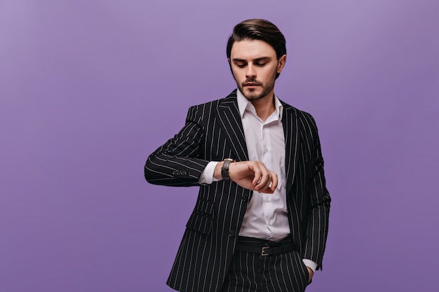 Kostenloses Foto stilvoller junger mann mit brünetten haaren, weißem hemd und klassischem gestreiftem anzug, der die uhr auf seiner hand isoliert über der violetten wand betrachtet