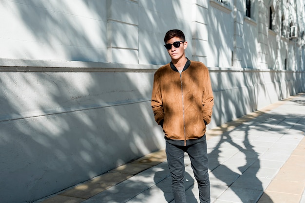 Stilvoller junger Teenager, der die braune Jacke und schwarze Sonnenbrille geht auf Bürgersteig trägt