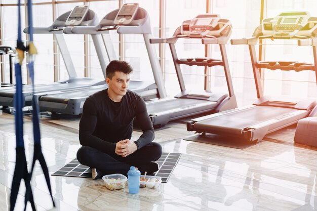 Stilvoller Kerl im Fitnessstudio, der sich auf dem Boden entspannt und gesundes Essen isst.