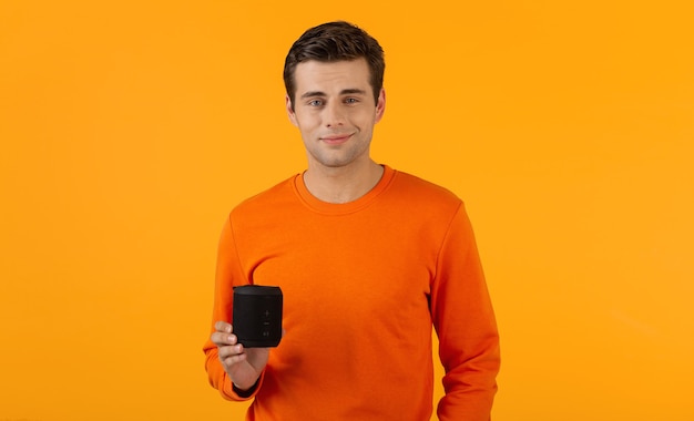 Stilvoller lächelnder junger mann im orangefarbenen pullover, der drahtlosen lautsprecher glücklich hört, musik zu hören, die spaß auf orange hat