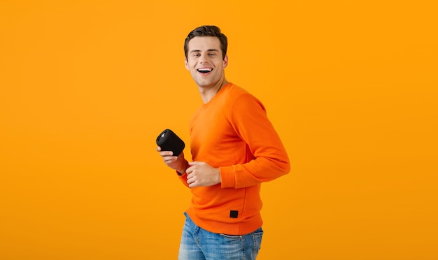 Stilvoller lächelnder junger Mann im orangefarbenen Pullover, der drahtlosen Lautsprecher glücklich hört, Musik zu hören, die Spaß auf Orange hat