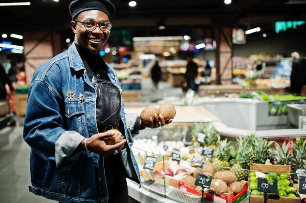 Stilvoller lässiger afroamerikanischer Mann in Jeansjacke und schwarzer Baskenmütze mit Kokosnüssen in der Bio-Obstabteilung des Supermarkts