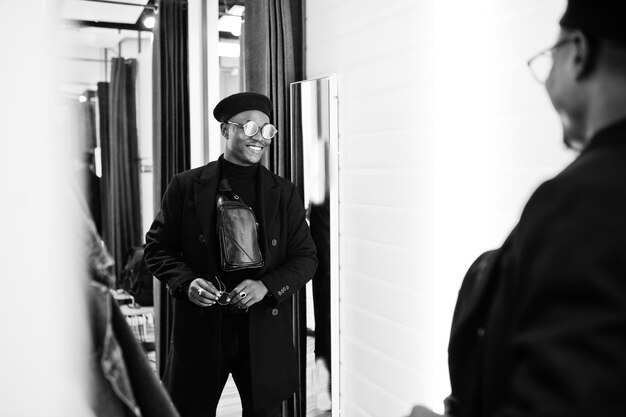 Stilvoller lässiger afroamerikanischer Mann mit schwarzer Baskenmütze und Mantel mit Gürteltasche im Umkleidekabinen-Bekleidungsgeschäft mit Blick auf den Spiegel