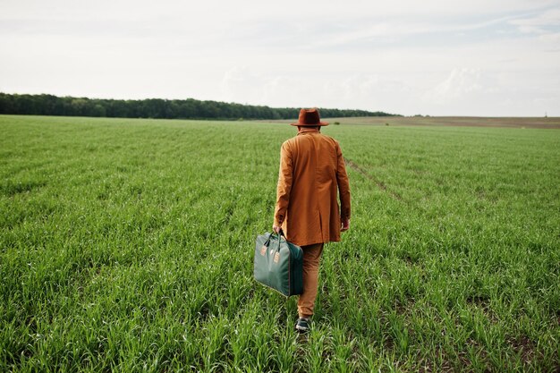 Stilvoller Mann in brillenbrauner Jacke und Hut mit Tasche auf grünem Feld