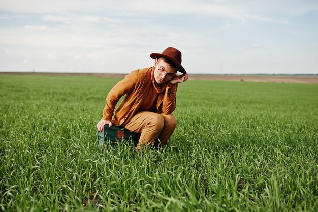 Stilvoller Mann in brillenbrauner Jacke und Hut mit Tasche auf grünem Feld