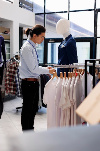 Stilvoller Mann überprüft Kleiderbügel mit weißen T-Shirts und arrangiert modische Kleidung vor der Ladeneröffnung. Mitarbeiter tragen formelle Kleidung und arbeiten mit trendigen Waren in einer modernen Boutique. Modekonzept