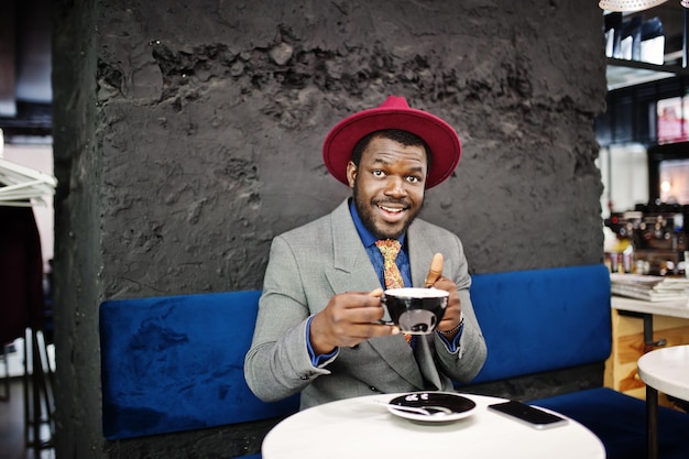 Stilvolles afroamerikanisches Mannmodell in grauer Jackenkrawatte und rotem Hut trinkt Kaffee im Café