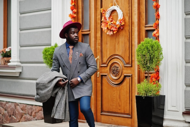Stilvolles afroamerikanisches Mannmodell in grauer Manteljacke, Krawatte und rotem Hut vor Herbststimmungstüren